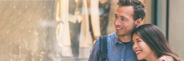 Casal multirracial fazendo compras juntos olhando para roupas na janela da loja andando na rua da cidade. Feliz mulher asiática abraçando marido homem caucasiano. Panorama banner fundo — Fotografia de Stock