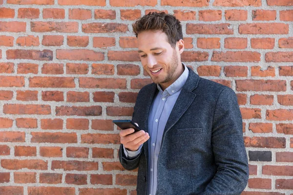 Young business man using smartphone in urban city background relaxing on brick wall texting sms on phone app living a modern lifestyle. Happy businessman playing mobile games — Stock fotografie