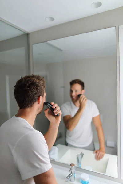 Homem barbear usando barbeador elétrico aparar sua barba em casa de banho- manhã grooming rotina pessoas conceito. Jovem olhando para o espelho se preparando. Cuidados faciais de beleza para homens — Fotografia de Stock