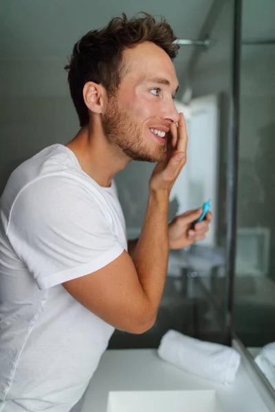 Man face cream beauty routine - facial skincare concept. Young adult in home bathroom looking at mirror putting moisturizer lotion in the morning taking care of dry skin. Moisturizing during winter — Stockfoto