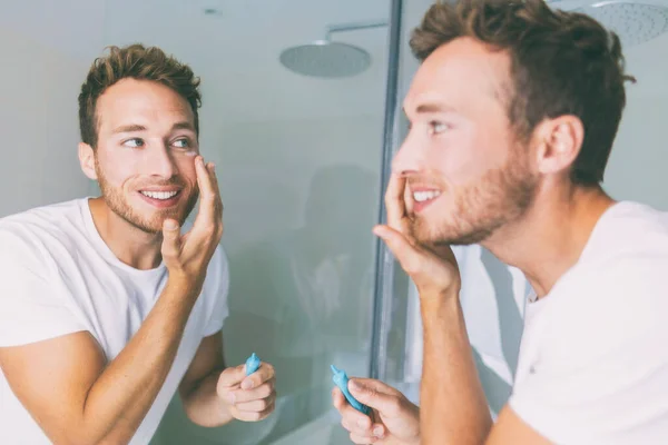 Man putting skincare facial treatment cream on face. Anti-aging skin care product. Male beauty morning routine at home lifestyle. Guy looking in bathroom mirror applying moisturizer under eyes — Stock Photo, Image