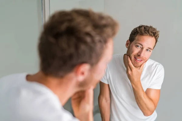Male beauty young man touching beard and face looking in mirror - healthy skin. Skincare in home bathroom. After shave men lifestyle shaving concept — Photo