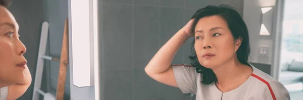 Madura mujer de mediana edad asiática tocándose el pelo mirándose a sí misma en el espejo del baño pancarta panorámica estilo de vida en casa. Colorante de cabello de señora china senior para cabello gris o peinado — Foto de Stock