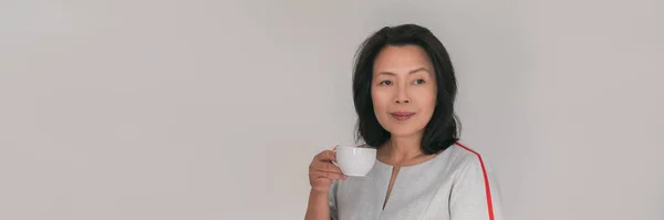 Olgun Asyalı kadın kahve fincanı içiyor. Kafede ya da ofiste dinleniyor. Panoramik afiş yapan iş adamlarının yaşam tarzına ara veriyor. 50 'li yaşlarda bir kadın. — Stok fotoğraf