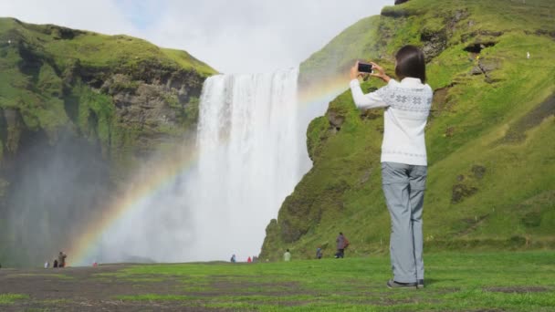 Vrouw toerist door waterval Skogafoss op IJsland het nemen van foto met telefoon — Stockvideo