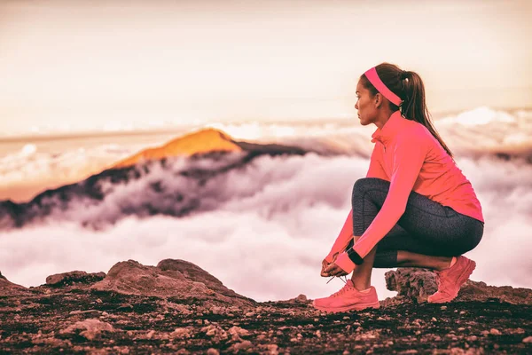 Atleta atando cordones de zapatillas de running con dispositivo de tecnología portátil reloj smartwatch para fitness y deportes. Sendero corredor mujer preparándose para correr en las montañas naturaleza paisaje en las nubes del atardecer —  Fotos de Stock