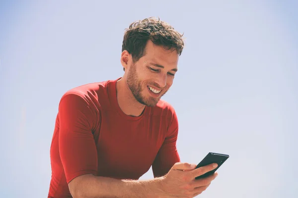 Gesunder Mann, der nach dem Training im Freien mit einer App auf seinem Mobiltelefon Videos anschaut. Lässiger junger männlicher Erwachsener Ende 20 im Sommer im Freien. Blauer Himmel Hintergrund. — Stockfoto