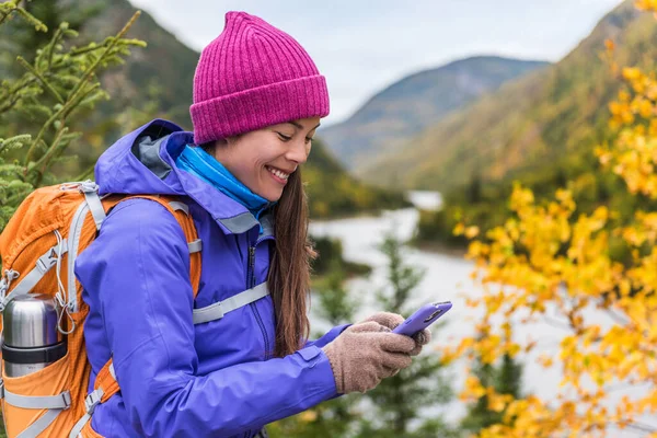 Happy asian girl in cold weather gear hiking in nature using smartphone with touch screen gloves during hike in autumn travel adventure in mountain forest outdoors. Beautiful landscape background. — Foto de Stock