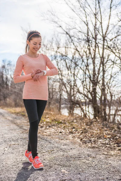 Active woman runner living a modern lifestyle getting ready for trail run on cold autumn forest path in morning nature outdoors. Running healthy girl happy of fitness routine looking at smartwatch. — Stock Photo, Image