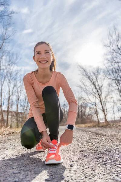 Autumn trail runner asian girl tying running shoes wearing sports smartwatch gadget gear. Female active athlete lacing shoe laces on forest path using smart watch heart rate fitness monitor. — Foto Stock