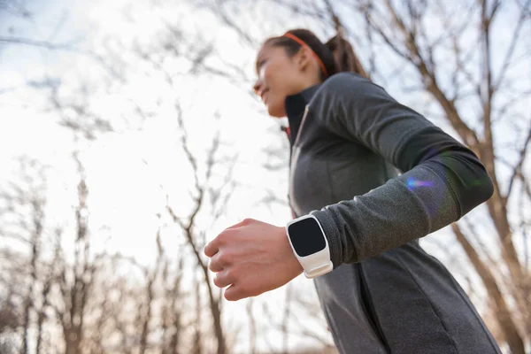 Runner wearing smartwatch on wrist closeup. Young fitness woman running working out cardio in autumn or winter nature outdoors on forest trail in sunlight fall wearing cold weather jacket. — Stock Photo, Image