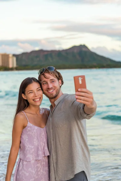 Para robi sobie selfie telefoniczne na plaży Waikiki o zachodzie słońca, Honolulu, na Hawajach. Młodzi ludzie na hawajskich wakacjach. Romantyczne miejsce wypoczynku na miesiąc miodowy — Zdjęcie stockowe
