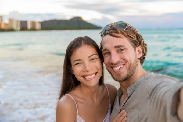 Selfie couple on Waikiki beach taking pictures with smartphone during night out walk on beach summer vacations in Honolulu, Hawaii. Travel destination. Young people having fun on hawaiian holidays — Foto Stock