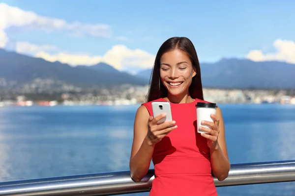 Businesswoman drinking coffee using mobile phone app in city to play video games or text sms online. Asian woman enjoying coffee break relaxing by watching smartphone content —  Fotos de Stock