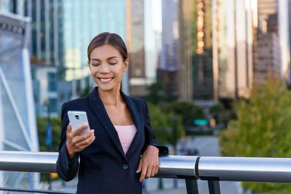 Žena sms sms pomocí aplikace na chytrém telefonu ve městě obchodní čtvrti. Mladá byznysmena, která používá smartphone, šťastně se usmívá, když má venku sako. Městská profesionálka ve věku 20 let — Stock fotografie