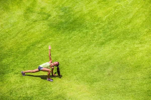 Läuferin streckt Beine beim Aufwärmen und macht einen Twist-Ausfallschritt auf dem grünen Rasenparkhintergrund. Glückliches Joggingmädchen trainiert im Sommer draußen — Stockfoto