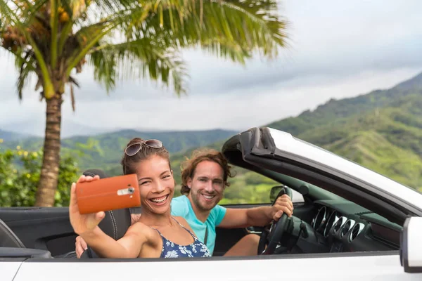 Car holiday selfie. Couple having fun on summer vacation road trip taking smartphone pictures during travel. Multiracial young people driving convertible — Foto Stock