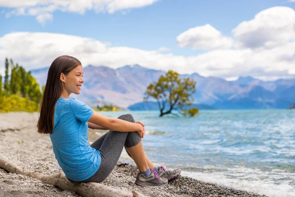 New Zealand Wanaka lake nature landscape travel woman traveler. Asian tourist relaxing enjoying view from beach shore at Wanaka lake landscape with lone tree, famous attraction. — Foto de Stock