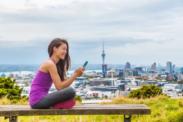 Auckland stad löpare flicka med hjälp av mobiltelefon app på skyline sommarpark. Vy över stadsbilden från Mount Eden, i Nordön, Nya Zeeland. Flicka tar paus utomhus efter löpning. — Stockfoto