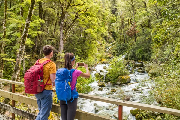 New zealand travel tourists taking phone picture on tramping hike in forest with backpacks. Woman holding smartphone taking photos of river on Routeburn track hikingtrail on South Island. — Stock Photo, Image