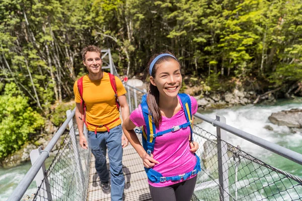 Nya Zeeland studsande par backpackers korsar flodbron. Asiatisk kvinna, vit man vandrar tillsammans med ryggsäckar på Routeburn Track Track trail path. — Stockfoto