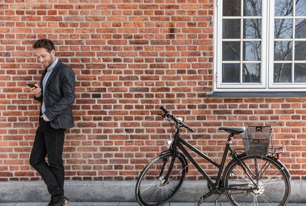 Young businessman going to work on bike commute using his mobile cellphone against city brick wall background. Happy business man bicycle commuter arriving at office using phone texting — Stock Photo, Image