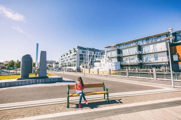 Reykjavik stad livsstil människor tittar på gatuscenen, Island. Kvinna med hjälp av smart telefon koppla av på bänk på Reykjavik hamn nära moderna bostadsbyggnad i centrum. Sommar Europa livsstil — Stockfoto