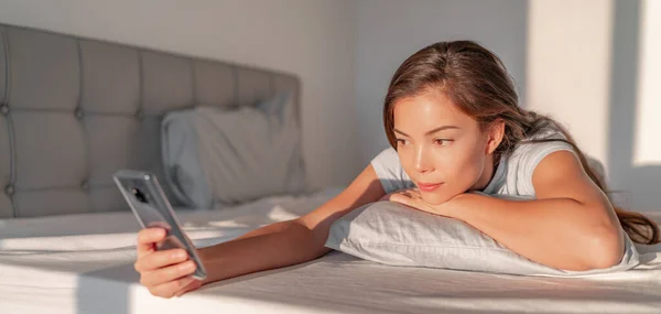 Mujer usando el teléfono en la cama mensajes de texto en el teléfono móvil después de despertarse por la mañana. Inicio estilo de vida gente banner panorama — Foto de Stock