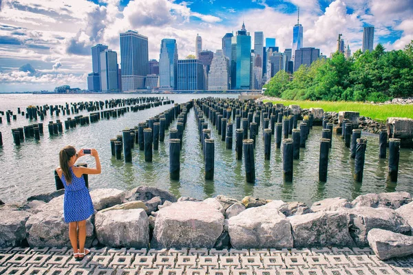 ニューヨークの観光客の女性はスマートフォンでモバイル写真を撮る。マンハッタンシティスカイラインウォーターフロントのライフスタイル。ブルックリン橋公園からのダウンタウンの景色を楽しむ人｜Pier 1塩沼 — ストック写真
