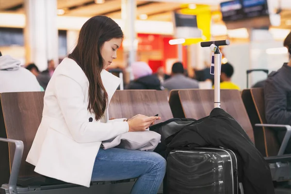 Pasajera del aeropuerto en el teléfono móvil a la espera de vuelo retrasado sentado en la puerta de la terminal con equipaje. persona aburrida y cansada —  Fotos de Stock