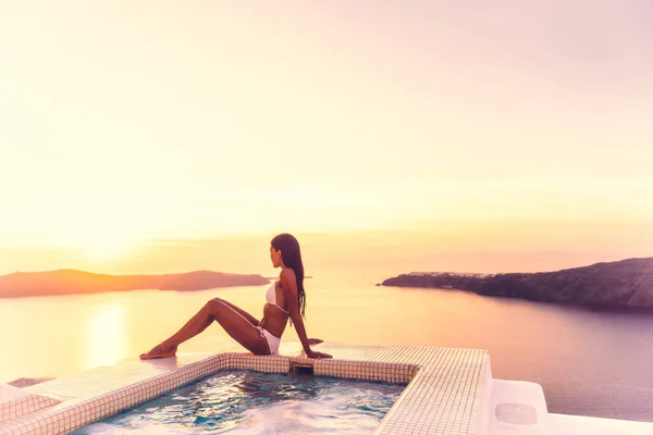 Luxury resort holiday bikini model woman relaxing lying down by hot tub jacuzzi piscina privada quarto do hotel suite varanda pôr do sol sobre o mar Egeu nas ilhas Cyclades, Santorini, Grécia. — Fotografia de Stock