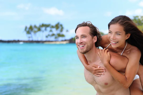 Beautiful healthy young people having fun. Multiracial piggyback couple on beach travel vacation in Caribbean destination. Happy smiling man piggybacking Asian woman — Stock Photo, Image