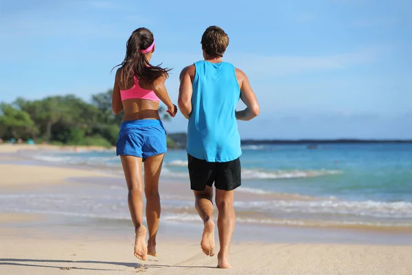 Zwei Fitness-Athleten laufen gemeinsam am Strand. Menschen von hinten joggen barfuß auf Sand an tropischen Reisezielen davon. Gesund fitte junge Erwachsene mit muskulösen schlanken Beinen trainieren das Herz-Kreislauf-System — Stockfoto