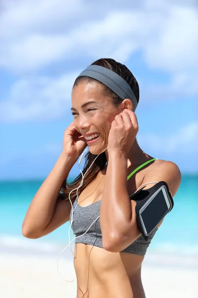 Runner girl wearing earphones and running armband phone arm strap getting ready for run workout. Gesture of putting on headset to listen to music while exercising on sunny beach summer vacation. — Stock Photo, Image
