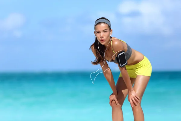 Müder atmender Läufer macht eine Laufpause am Strand mit Meereshintergrund. Asiatische chinesische Athletin ruht die Hände auf den Knien und nimmt erschöpft eine Pause nach dem Training ein, um Luft zu holen. — Stockfoto