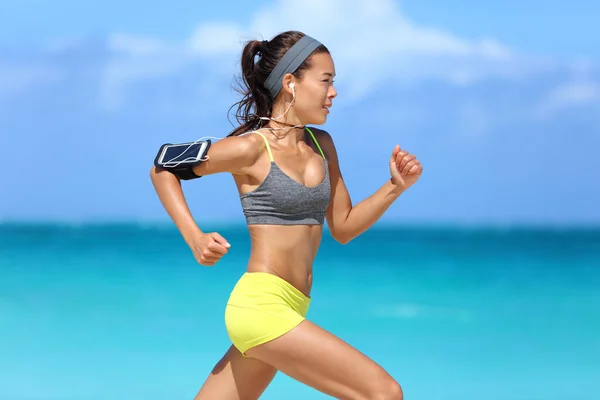 Atleta corriendo corredor mujer escuchando música en su brazalete deportivo teléfono con pantalla táctil y auriculares auriculares en la playa de verano. Chica fitness trotar entrenamiento rápido cardio y glúteos. —  Fotos de Stock