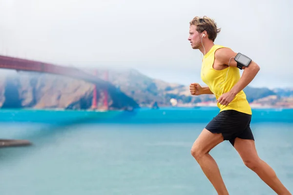 Un coureur écoutant de la musique pendant l'entraînement cardio. Athlète en cours d'exécution en utilisant l'application sur le brassard smartphone et écouteurs. Jeune jogging sportif près de la baie de San Francisco et du Golden Gate Bridge. — Photo