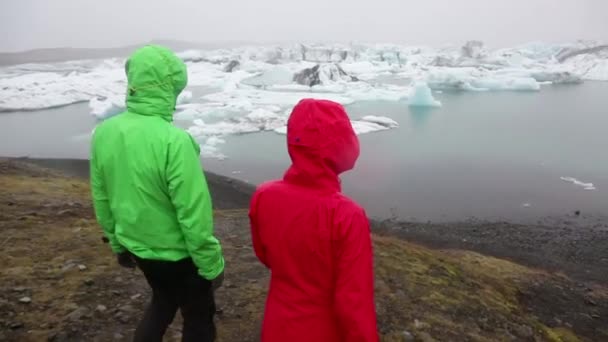 Прогулочная пара Исландии на ледниковом озере Джоколсарлон ходит в бронежилетах под дождем. Активный образ жизни туристов прогулки наслаждаясь красивой исландской природы — стоковое видео