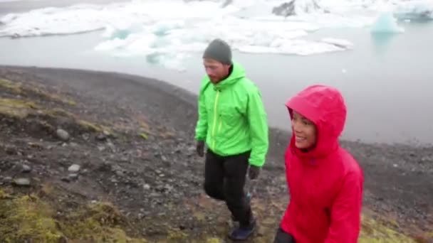 Mensen met zware regenval. IJslandse wandelpaar door Jokulsarlon gletsjermeer wandelen in hardshell jassen in de regen. Actieve levensstijl mensen wandelen genieten van prachtige IJslandse natuur — Stockvideo