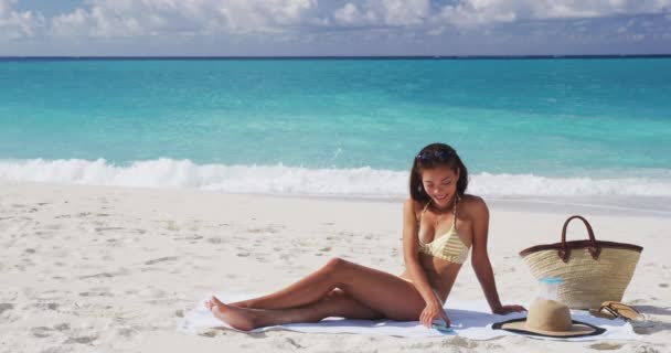 夏天旅行时，女人在海滩上使用手机应用程序，在沙滩上晒日光浴。夏天，穿着比基尼躺在沙滩毛巾上的年轻女性穿着比基尼在海上悠闲自在地躺着 — 图库视频影像