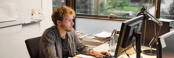 若い博士課程の学生は大学で試験のための最終論文を書いて勉強します.図書室のデスクトップコンピュータに入力するオフィスのデスクで働く男 — ストック写真