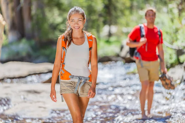 Мандрівники ходять лісом через струмок води, тримаючи чоботи, які ходять по природі Йосеміті. Щаслива азіатська дівчинка і чоловік влітку мандрують з рюкзаками — стокове фото