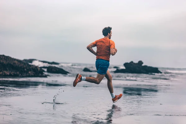 Ejercicio hombre atleta al aire libre corriendo sobre arena mojada en el entrenamiento de playa cardio corriendo rápido. Perfil del corredor en ropa deportiva jogging — Foto de Stock