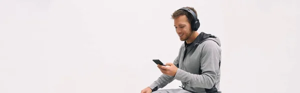 自宅の白い壁の背景のパノラマに座ってヘッドフォンを身に着けている音楽携帯電話アプリを聞いて男。ハッピー男アクティブライフスタイルスポーツ選手使用スマートフォンでジム外パノラマバナー — ストック写真