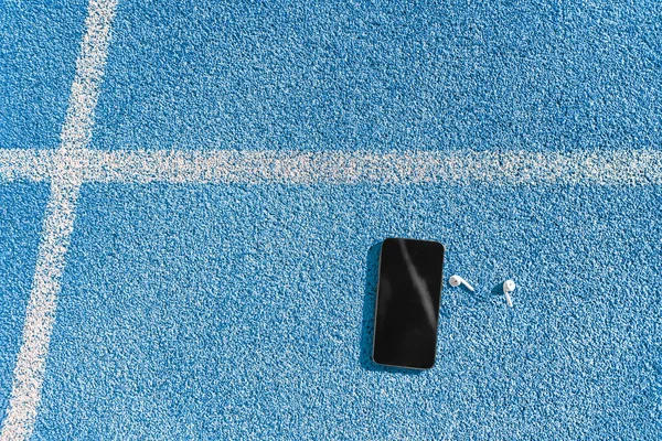 Trilha de corrida e pista de campo e smartphone para ouvir música no aplicativo de telefone móvel com fones de ouvido. Textura de fundo azul de pistas pistas para copyspace para conceitos de fitness e esportes — Fotografia de Stock