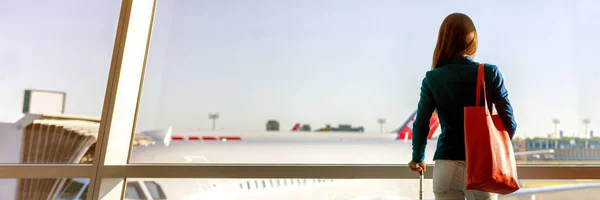 Panoramiczne tło podróży lotniczej kobiety turystki patrzącej na samoloty okienne na pasie startowym z podręcznym banerem bagażowym. Pierwsza klasa podróży służbowej salon czeka na pokład lotu przy wylocie bramki — Zdjęcie stockowe