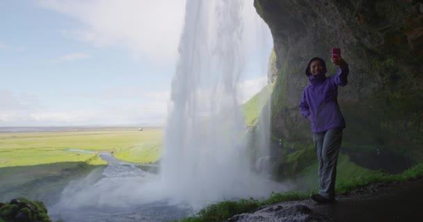 アイスランドの女性観光客は滝のそばでスマートフォンを使って自撮り写真を撮る — ストック動画