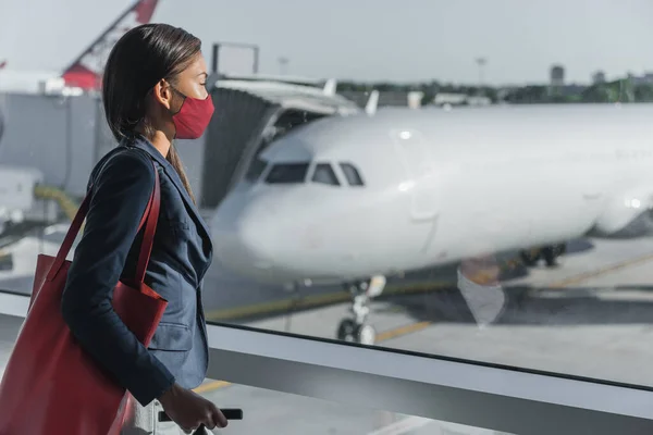 Travel by plane during coronavirus. Asian tourist waiting for flight at terminal wearing face mask indoors looking at window with airplane on tarmac — Stock Photo, Image