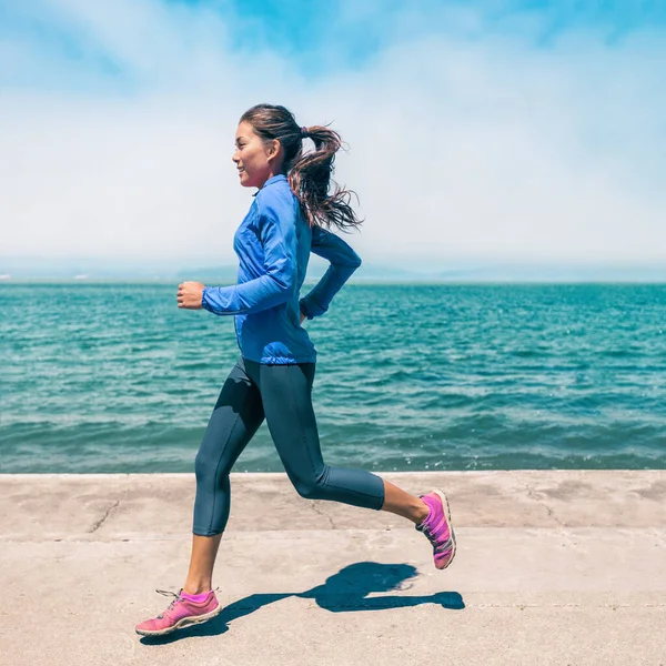 Running idrottsman asiatisk kvinna jogging vid stranden bär blå windbreaker jacka leggings och skor. Träning utomhus på våren vid havet. Porträtt av fyrkantiga grödor. — Stockfoto