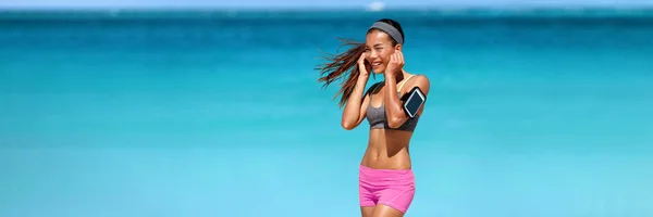 Lyssnar på podcast på telefon app med trådlösa hörlurar under beach workout. Passar asiatisk löpare kvinna glad med bärbar teknik armband utanför banner panoramautsikt. — Stockfoto
